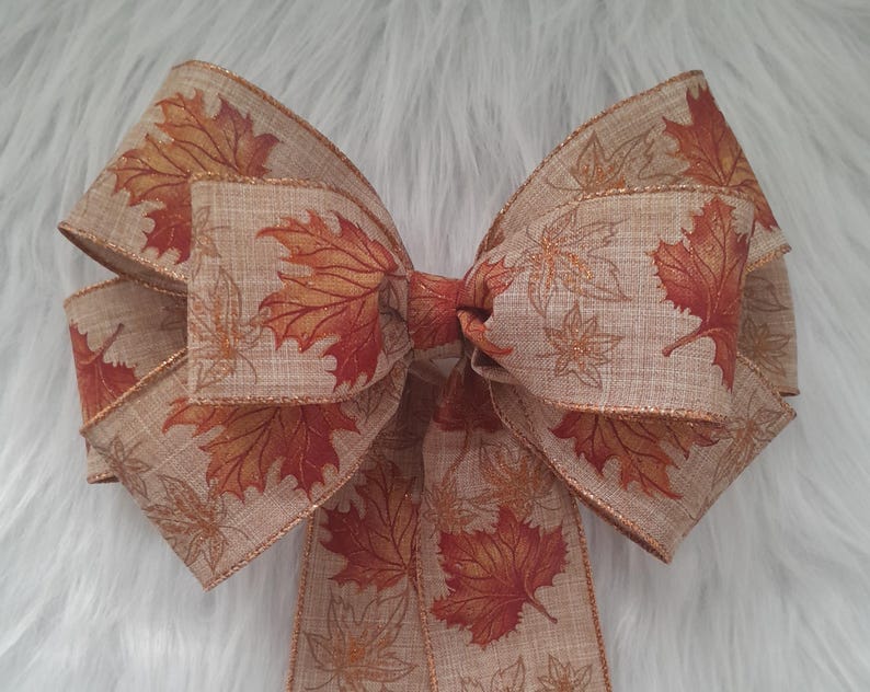 May include: A decorative bow featuring a tan fabric with a leaf pattern in shades of orange and red. The bow has a gold glitter trim and is tied in the center. The bow is likely used for seasonal decor, such as autumn or fall.