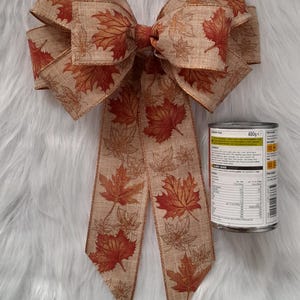 May include: A large decorative bow with a fall leaf pattern in shades of brown, orange, and red. The bow is made of fabric and has long streamers. A can of food is next to the bow.