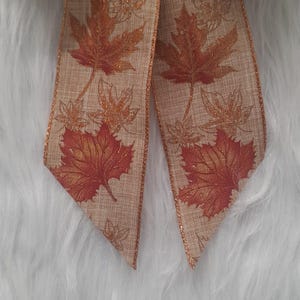 May include: Close-up of a decorative ribbon featuring a fall leaf design. The ribbon has a tan background with large, detailed orange and red maple leaves. Smaller, gold-colored leaves are also printed on the ribbon.