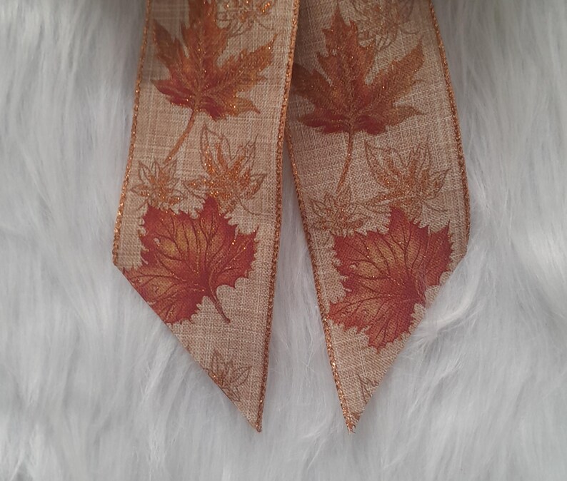May include: Close-up of a decorative ribbon featuring a fall leaf design. The ribbon has a tan background with large, detailed orange and red maple leaves. Smaller, gold-colored leaves are also printed on the ribbon.