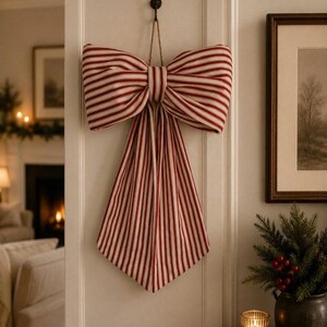 May include: A large decorative bow made of fabric with a red and white vertical striped pattern. The bow features two wide loops and two long, pointed tails hanging downward, suspended from a dark hook by a thin, natural-colored twine loop.