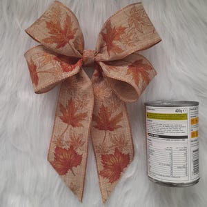 May include: A decorative bow with a fall theme. The bow is made of tan ribbon with a maple leaf pattern in shades of orange and brown. The bow is next to a can of food.