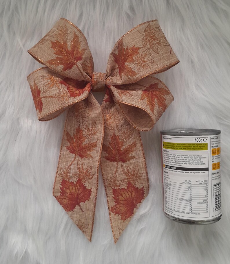 May include: A decorative bow with a fall theme. The bow is made of tan ribbon with a maple leaf pattern in shades of orange and brown. The bow is next to a can of food.