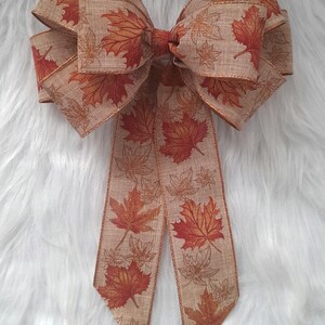 May include: A large decorative bow featuring a fall leaf pattern. The bow is made of a tan-colored fabric with a repeating design of red and orange maple leaves. The edges are trimmed with a gold metallic thread.