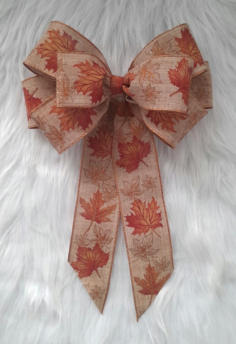 May include: A large decorative bow featuring a fall leaf pattern. The bow is made of a tan-colored fabric with a repeating design of red and orange maple leaves. The edges are trimmed with a gold metallic thread.