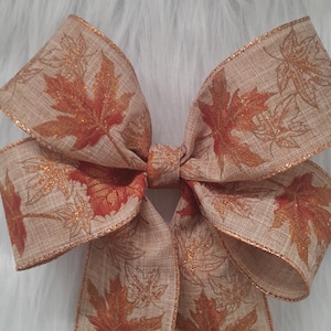 May include: A large decorative bow with a fall theme. The bow is made of tan fabric with a maple leaf pattern in shades of orange and brown. The edges are trimmed with gold glitter.