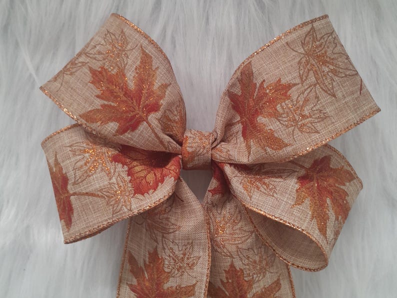 May include: A large decorative bow with a fall theme. The bow is made of tan fabric with a maple leaf pattern in shades of orange and brown. The edges are trimmed with gold glitter.