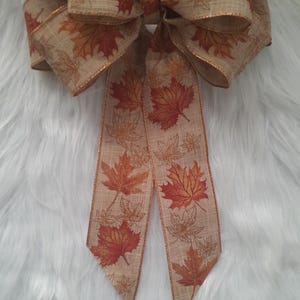 May include: A large decorative bow featuring a fall leaf design. The bow is made of tan fabric with a repeating pattern of orange and brown maple leaves. The edges are trimmed with a metallic gold ribbon.