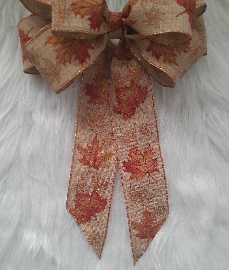 May include: A large decorative bow featuring a fall leaf design. The bow is made of tan fabric with a repeating pattern of orange and brown maple leaves. The edges are trimmed with a metallic gold ribbon.