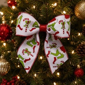 May include: A decorative bow featuring a white fabric base with a repeating pattern of green legs wearing red shoes and red and white striped candy canes. The centre of the bow is made of red glittery material, and the edges are finished with red stitching.