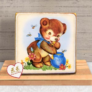 May include: A square wooden block featuring a vintage illustration of a brown teddy bear with a blue bow, eating honey from a blue pot. Bees and flowers surround the bear. The block is on a wooden surface, with a heart-shaped logo in the corner.