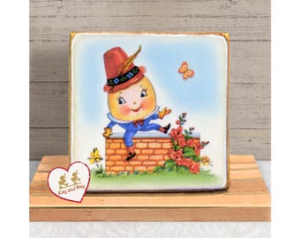 May include: A square wooden block featuring a vintage illustration of Humpty Dumpty. The character is depicted with a smiling face, wearing a red hat and blue outfit, sitting on a brick wall with flowers and butterflies. The block is displayed on a wooden surface.