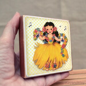 May include: A square wooden block featuring a vintage illustration of a hula dancer in a yellow skirt and floral leis. The background is yellow with white polka dots. Musical notes are scattered around the dancer. The block is held in a hand.