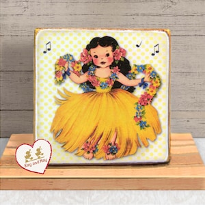 May include: A decorative wooden block featuring a vintage illustration of a hula dancer in a yellow skirt and floral lei. The background is dotted with yellow circles. Musical notes are in the background. The block is on a wooden stand. The text "Kay and May" is on a heart-shaped sticker.