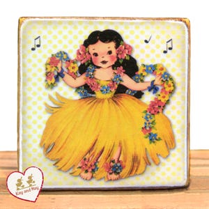 May include: Wooden block featuring a vintage illustration of a hula dancer in a yellow dress, adorned with floral leis and hair flowers. The background is a yellow polka dot pattern with musical notes. The block has a heart-shaped logo that says "Kay and May".