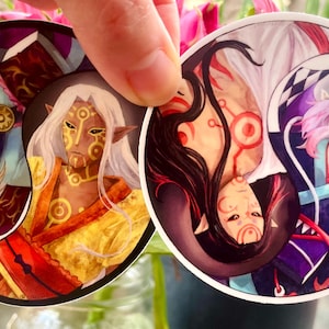 Può includere: Due adesivi rotondi con personaggi in stile anime in un design yin-yang. Un adesivo presenta un personaggio con capelli bianchi e dettagli dorati, l'altro ha un personaggio con capelli rosa e un berretto blu scuro. Gli adesivi sono tenuti da una mano.