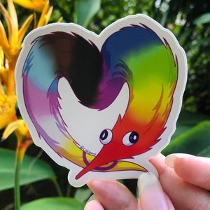 May include: A heart-shaped sticker featuring a colorful worm-like creature. The worm has a red head, a rainbow-colored body, and large, cartoonish eyes. The sticker has a white border and the artist's name, "@rocyart", is printed on the sticker.