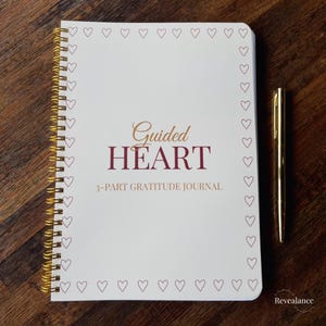 May include: A white spiral-bound journal with the words "Guided Heart" and "3-PART GRATITUDE JOURNAL" in burgundy and gold lettering. The journal is decorated with small heart outlines and a gold pen rests beside it.