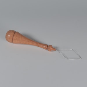 May include: A wooden needle threader with a light brown handle and a diamond-shaped wire loop. The handle has a bulbous top and a tapered body, with a small decorative detail near the wire loop. The tool is designed for threading needles.