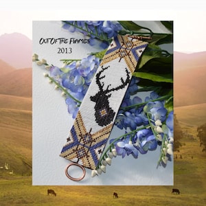 May include: A white beaded bracelet with a black silhouette of a deer with antlers. The bracelet has gold and blue accents and a copper clasp. The bracelet is on a background of blue flowers.