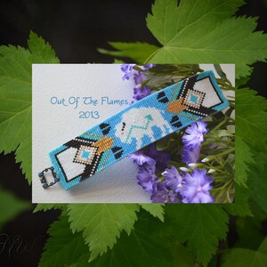May include: A beaded bracelet with a white bison design on a blue background. The bracelet has black, gold, and turquoise accents. The text "Out Of The Flames 2013" is visible on the white background.