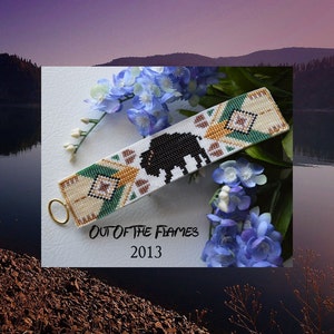 May include: A beaded bracelet with a white background, featuring a black buffalo design and geometric patterns in tan, green, and cream. The bracelet has a gold-colored ring and the text "OUT OF THE FLAMES 2013".