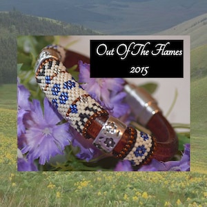 Puede incluir: Un brazalete de cuero marrón con detalles plateados. Las cuentas son blancas con detalles azules y marrones. El brazalete está sobre un lecho de flores moradas. El texto "Out Of The Flames 2015" está en el fondo.
