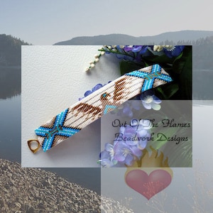 May include: A white beaded bracelet with blue and brown accents. The bracelet features a geometric design with a star shape and a bird design. The bracelet has a metal clasp. The text "Out of the Flames Beadwork Designs" is visible in the image.