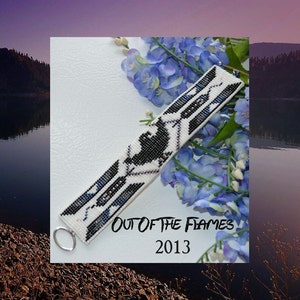 Puede incluir: Un brazalete blanco de cuentas con patrones geométricos negros y azules. El brazalete tiene un cierre plateado y está rodeado de flores azules. El texto "OUT OF THE FLAMES 2013" está impreso debajo del brazalete.