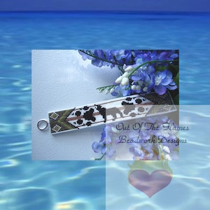 May include: A white beaded bracelet with black and brown geometric patterns. The bracelet has a silver clasp and is displayed against a blue background with flowers.