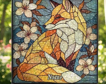 Könnte beinhalten: Buntglasfenster mit einem Fuchs und floralem Design. Der Fuchs ist in Orange-, Gelb- und Weißtönen dargestellt, umgeben von weißen Blüten und braunen Blättern auf blauem Hintergrund. Das Panel ist schwarz gerahmt und hängt an einer Kette.