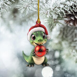 May include: A green alligator wearing a red Santa hat holds a red and white Christmas ornament. The ornament has a snowflake design.