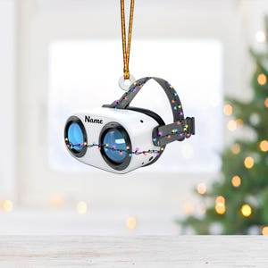 May include: White virtual reality headset ornament with a string of colorful lights wrapped around it. The headset has a black strap and says "Name" on the front.