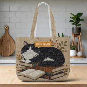 May include: A beige tote bag featuring a black and white cat lying on a stack of books. The background has a subtle tan floral pattern. A rectangular orange label with the text "YOUR NAME" is positioned above the cat. The bag has two handles.