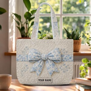 May include: A quilted tote bag in cream with a blue floral pattern, featuring a large central bow and a rectangular patch at the bottom with the text YOUR NAME. The bag has two matching shoulder straps and a textured, stitched surface design.