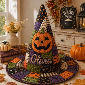 May include: A quilted witch hat featuring a patchwork design of orange, black, purple, and green fabrics with patterns of stars, spiderwebs, and bats. The hat displays a large orange jack-o'-lantern appliqué and the embroidered name Olivia.