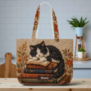 May include: A quilted tote bag featuring a black and white cat sleeping on a stack of books. The bag has floral patterned straps and a beige background with orange flowers. The word Name is printed on a rectangular label on the bottom book.