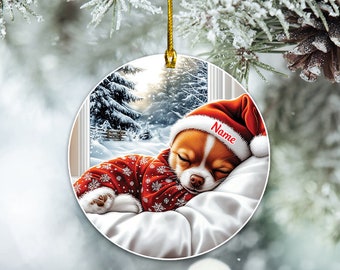 Könnte beinhalten: Ein weißer Keramik-Weihnachtsbaumschmuck mit einem schlafenden Chihuahua, der eine rote Weihnachtsmütze und einen Pyjama mit Schneeflocken trägt. Der Anhänger hat eine Fensterszene im Hintergrund mit einem verschneiten Wald. Der Text "Name" steht auf der Mütze.