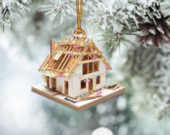 May include: A wooden ornament shaped like a house under construction with a string of colourful lights. The ornament is hanging from a gold string and has the word "Name" written on the base.