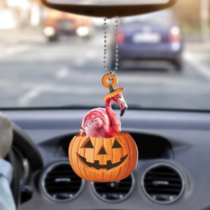 May include: A pink flamingo with a pumpkin hat hanging from a rearview mirror. The flamingo is inside a carved orange pumpkin with a jack-o'-lantern face.