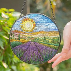May include: A stained glass suncatcher depicting a lavender field with a bright yellow sun in a blue sky. The suncatcher is round and has a black metal frame.