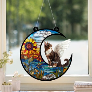 May include: A stained glass window hanging featuring a crescent moon with a cat with wings sitting on it. The moon is surrounded by sunflowers and other flowers. The cat is brown and white and has a name tag that says "Name".