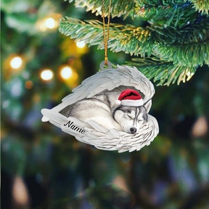 May include: A white ceramic Christmas ornament featuring a sleeping husky dog wearing a red Santa hat. The dog is nestled inside a pair of white angel wings. The ornament has the word "Name" inscribed on it.