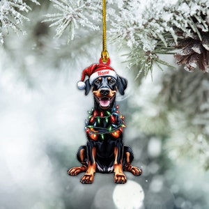 May include: A black and tan Doberman Pinscher wearing a red Santa hat and a string of colorful Christmas lights around its neck. The ornament is ready to hang on a Christmas tree.