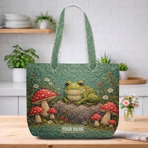 May include: A quilted tote bag featuring a green background with a printed illustration of a frog sitting on a rock surrounded by red and white mushrooms and small white flowers. The bottom center includes a rectangular label with the text YOUR NAME.