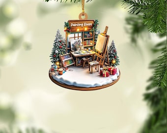 Könnte beinhalten: Ein hölzerner Weihnachtsbaumschmuck in Form eines Miniatur-Malerzimmers mit einem Schild, auf dem "Painting Room" steht. Der Anhänger zeigt eine kleine Staffelei, Pinsel und einen Tisch mit Farbtuben und einer Palette. Auf beiden Seiten des Raumes stehen zwei kleine Weihnachtsbäume.
