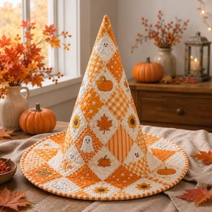 May include: A quilted witch hat featuring a diamond-patterned patchwork design in shades of orange, white, and yellow. The fabric displays embroidered motifs of ghosts, pumpkins, sunflowers, and autumn leaves, finished with a stitched brim edge.