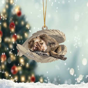 May include: A brown monkey with closed eyes is sleeping inside a white angel wing ornament. The ornament has the name "Bella" written on it.
