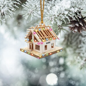 May include: A wooden house ornament with a roof and string lights. The ornament has the word "NAME" on the side. The ornament is hanging from a gold string. The background is a blurred image of a snowy tree.