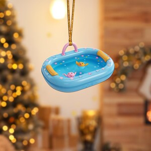 May include: A blue, oval-shaped ornament resembling a kiddie pool, with a pink handle and two yellow and pink figures swimming inside. The pool is decorated with pink stars and yellow accents. It hangs from a gold string.